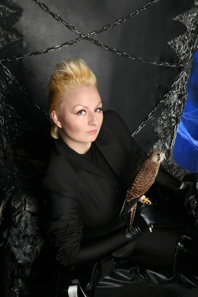 Foto de uma fêmea bruxa rainha segurando pássaro e sentado em um trono negro assustador gótico — Fotografia de Stock