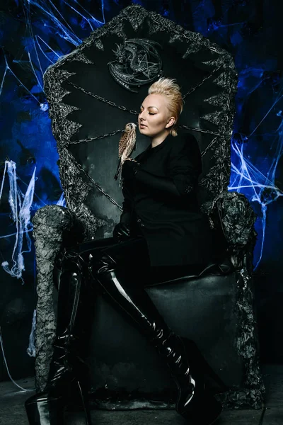 女性魔女の女王が鳥を抱いてゴシック様式の恐ろしい黒い玉座に座っている写真 — ストック写真
