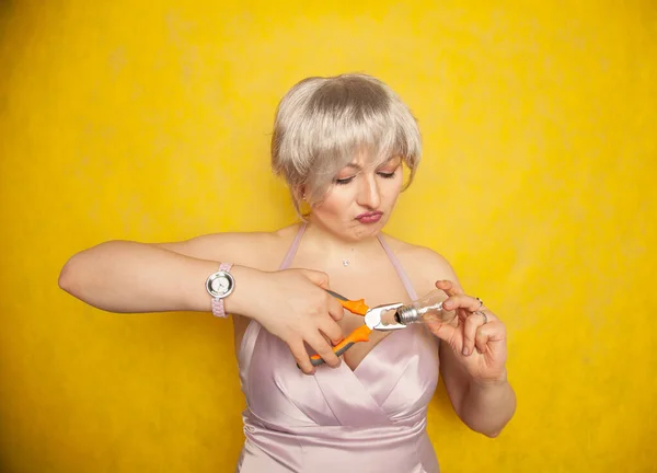 The blonde girl does not know how to make repairs. a confused woman stands with construction tongs and a light bulb. — Stock Photo, Image