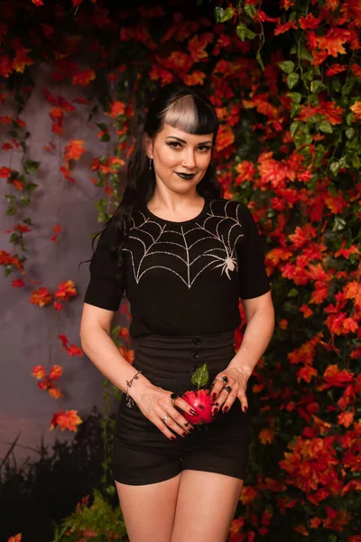 Schöne Halloween-Mädchen in schwarzer Pin-up-Kleidung mit totem roten Apfel in der Hand auf Herbstblättern Hintergrund allein — Stockfoto