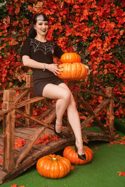 Sexy y atractiva bruja morena en otoño colorido parque con hojas naranjas y rojas posando con calabazas —  Fotos de Stock