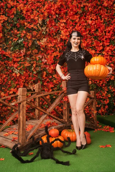 Sexy e atraente bruxa morena no outono colorido parque com laranja e folhas vermelhas posando com abóboras — Fotografia de Stock