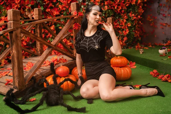 Linda chica sexy pin up en traje negro relajarse sentado en la hierba con el fondo de las hojas de otoño. negro cabello bruja mujer comer halloween caramelo . —  Fotos de Stock