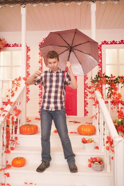 Adolescente en otoño. Retrato de un buen adolescente caminando cerca de la casa con un paraguas en un cálido día de otoño. niño divertirse al aire libre durante el mal tiempo en la lluvia —  Fotos de Stock