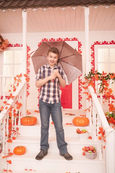 Adolescente in autunno. Ritratto di un bel ragazzo che cammina vicino alla casa con un ombrello in una calda giornata autunnale. bambino divertirsi all'aperto durante il brutto tempo sotto la pioggia — Foto Stock