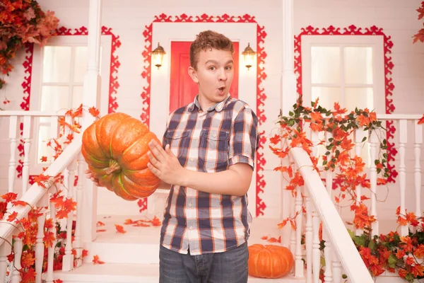 Happy young person with pumpkin on autumn background with leaves. teenager boy in a plaid cozy shirt on the decorated porch of the white house