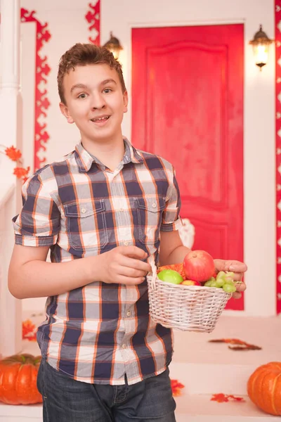 Un joven sosteniendo una cesta de fruta. un adolescente en una camisa acogedora a cuadros en el porche decorado de la casa blanca en el otoño —  Fotos de Stock