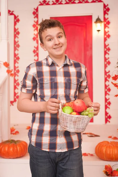 Un joven sosteniendo una cesta de fruta. un adolescente en una camisa acogedora a cuadros en el porche decorado de la casa blanca en el otoño —  Fotos de Stock