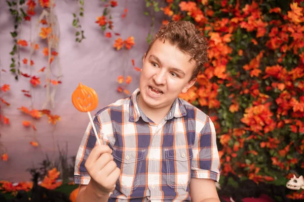 Boos ontevreden tiener in geruite shirt zit met pompoen Lollipop en toont een kwaad gezicht tegen de achtergrond van de herfst bladeren. het kind is ontevreden met het resultaat van truc of traktatie — Stockfoto