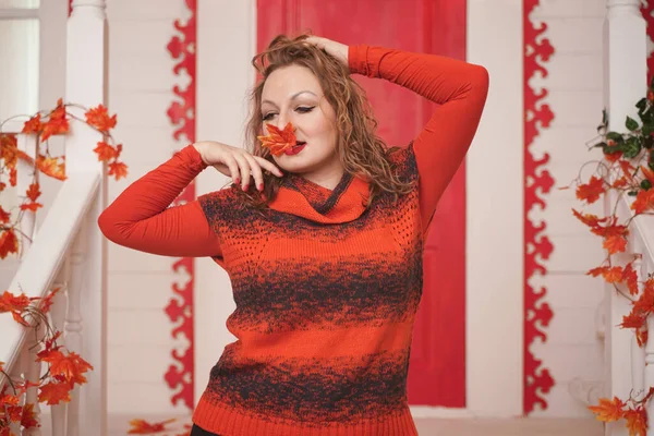 Más tamaño chica caucásica en suéter naranja sosteniendo otoño hoja de arce rojo en la mano con la cara cerrada. chica adulta se para en el porche de su casa blanca . — Foto de Stock
