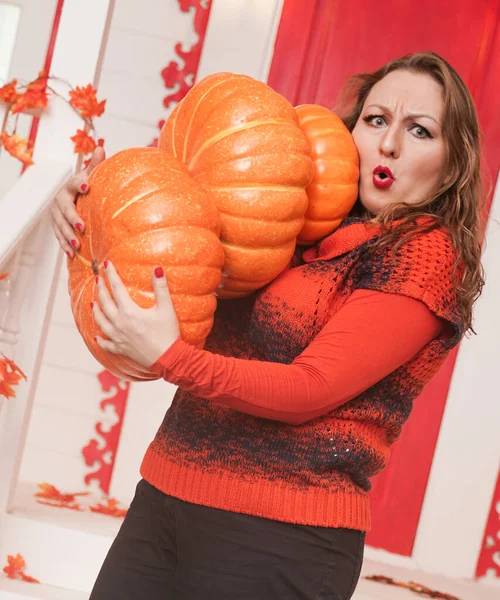 Joven mujer caucásica gordita en suéter naranja sosteniendo calabazas de pie en el porche de su casa blanca . — Foto de Stock