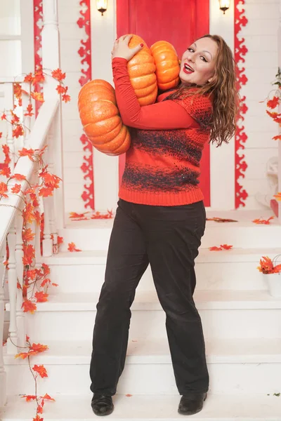 Joven mujer caucásica gordita en suéter naranja sosteniendo calabazas de pie en el porche de su casa blanca . — Foto de Stock