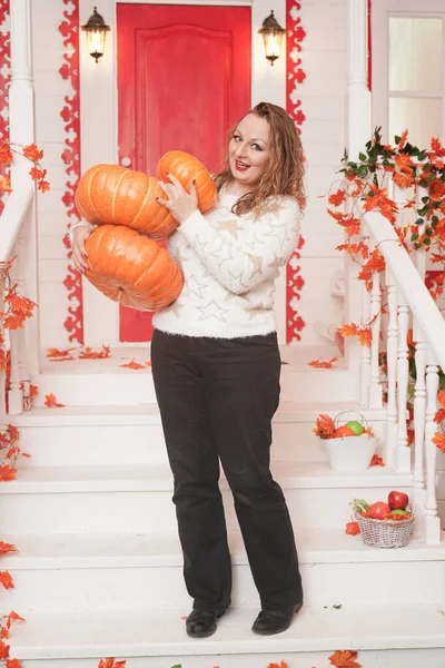 Adulte mignonne grosse fille dans un pull blanc avec des étoiles d'or tient une pile de grandes citrouilles et se tient sur le porche de sa maison blanche . — Photo