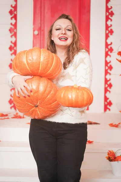 Adulto linda chica gorda en un suéter blanco con estrellas de oro sostiene una pila de calabazas grandes y se encuentra en el porche de su casa blanca . — Foto de Stock