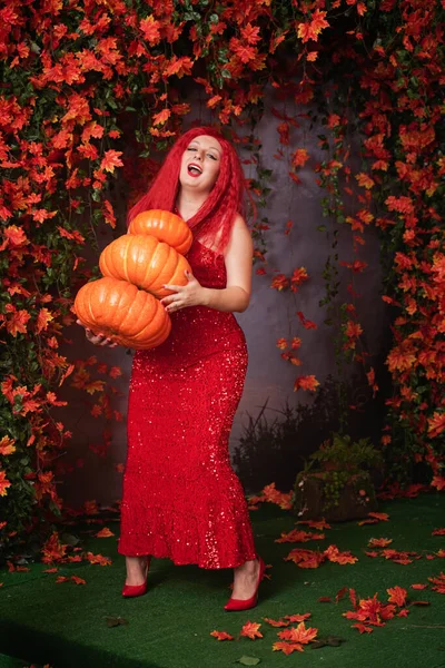 빨간 긴 옷을 입고 시퀀스를 걸친 다 자란 귀엽고 작은 여자 아이는 커다란 호박을 쌓아 놓고 가을 오렌지 잎 사이에 있는 풀 위에 서 있습니다 — 스톡 사진