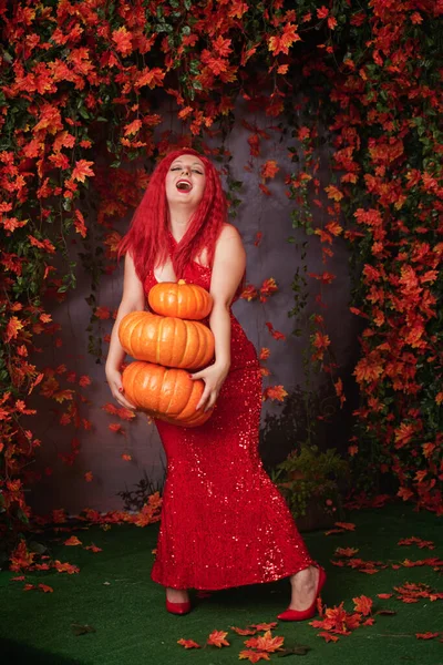 Adulto lindo más chica de tamaño en un vestido largo rojo con lentejuelas sostiene una pila de calabazas grandes y se encuentra en la hierba entre el follaje naranja de otoño —  Fotos de Stock
