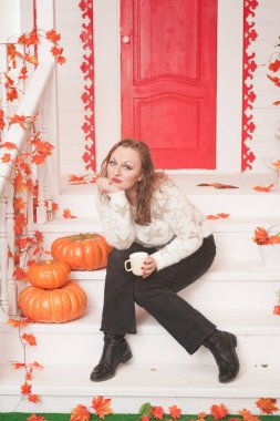 Güzel, iri yarı, beyaz kazaklı bir kadın günün ilk kahvesini içiyor. Kırmızı kapılı, güzel bir sonbahar evinin verandasında oturuyor.