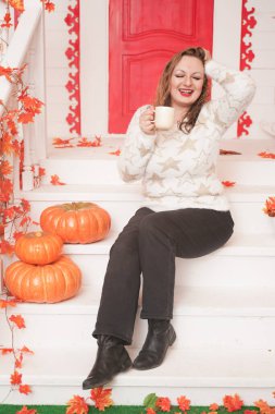 Güzel, iri yarı, beyaz kazaklı bir kadın günün ilk kahvesini içiyor. Kırmızı kapılı, güzel bir sonbahar evinin verandasında oturuyor.