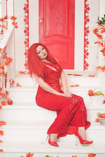 Young stylish chubby woman wearing red maxi dress on the porch of her white house. Fall fashion, elegant look. Plus size model.