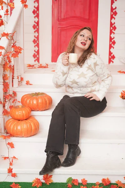 Muito jovem mulher plus size em camisola branca acolhedora bebendo o primeiro café do dia sentado no alpendre de uma bela casa de outono com uma porta vermelha — Fotografia de Stock