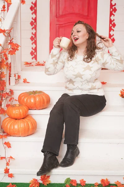 Красивая молодая женщина plus size в уютном белом свитере, пьющая первый кофе в день, сидя на крыльце красивого осеннего дома с красной дверью — стоковое фото