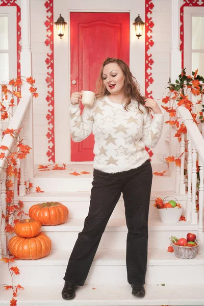 Bastante joven mujer de tamaño grande en suéter blanco acogedor bebiendo el primer café del día sentado en el porche de una hermosa casa de otoño con una puerta roja — Foto de Stock