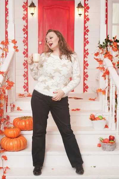 Bastante joven mujer de tamaño grande en suéter blanco acogedor bebiendo el primer café del día sentado en el porche de una hermosa casa de otoño con una puerta roja — Foto de Stock