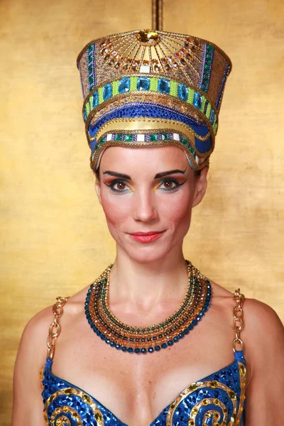 Beautiful thin Egyptian woman with pronounced cheekbones in a huge gold headdress without hair in a blue swimsuit on the background in the Studio