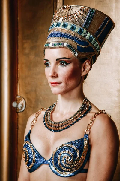 Mulher egípcia fina bonita com maçãs do rosto pronunciadas em uma enorme cobertura para a cabeça de ouro sem cabelo em um maiô azul no fundo no estúdio — Fotografia de Stock