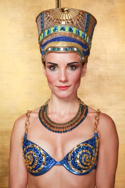 Beautiful thin Egyptian woman with pronounced cheekbones in a huge gold headdress without hair in a blue swimsuit on the background in the Studio