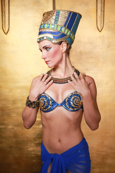Beautiful thin Egyptian woman with pronounced cheekbones in a huge gold headdress without hair in a blue swimsuit on the background in the Studio — Stock Photo, Image