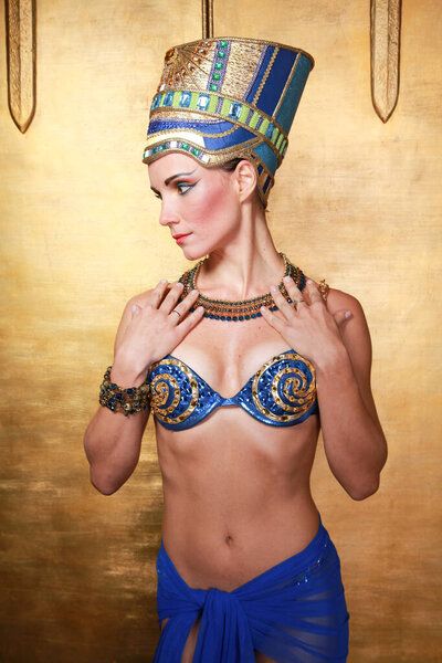 Beautiful thin Egyptian woman with pronounced cheekbones in a huge gold headdress without hair in a blue swimsuit on the background in the Studio