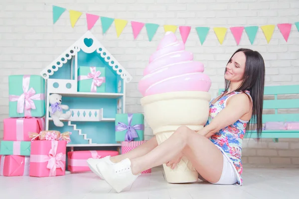 Mooie fitness meisje in witte shorts en stijlvolle korte jurk poseren met enorme kunstmatige voedsel desserts op de achtergrond van de kamer. Charmante vrolijke slanke vrouw spelen met voedsel tijdens het dieet. — Stockfoto