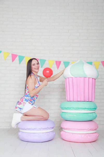 Mooie fitness meisje in witte shorts en stijlvolle korte jurk poseren met enorme kunstmatige voedsel desserts op de achtergrond van de kamer. Charmante vrolijke slanke vrouw spelen met voedsel tijdens het dieet. — Stockfoto