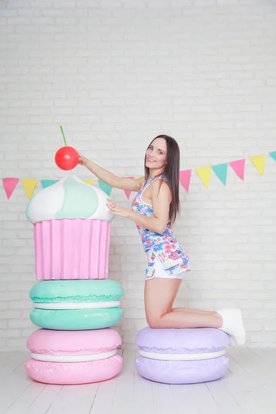 Beautiful fitness girl in white shorts and stylish short dress posing with huge artificial food desserts on the background of the room. Charming cheerful slender woman playing with food while dieting.