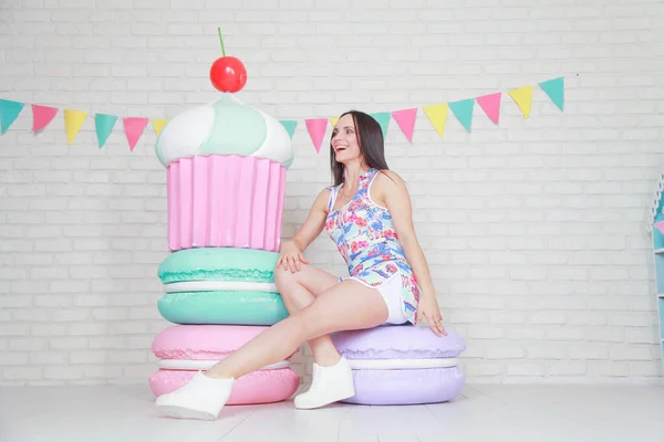 Mooie fitness meisje in witte shorts en stijlvolle korte jurk poseren met enorme kunstmatige voedsel desserts op de achtergrond van de kamer. Charmante vrolijke slanke vrouw spelen met voedsel tijdens het dieet. — Stockfoto