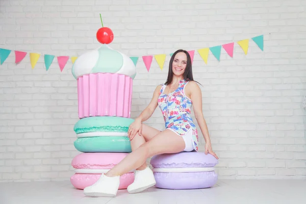 Mooie fitness meisje in witte shorts en stijlvolle korte jurk poseren met enorme kunstmatige voedsel desserts op de achtergrond van de kamer. Charmante vrolijke slanke vrouw spelen met voedsel tijdens het dieet. — Stockfoto