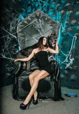Küçük siyah elbiseli güzel, zayıf bir kadın. Büyük Danimarkalı köpeğiyle birlikte Cadılar Bayramı 'nın arka planında büyük, korkunç siyah tahtta poz veriyor.