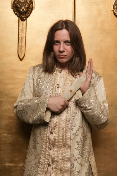 Charmante schüchterne junge Teenager in einem mittelalterlichen goldenen Anzug in einem Fotostudio ist bereit, auf eine Party zu gehen. ein Mann mit einem Messer aus Jade — Stockfoto