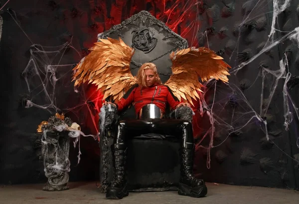 Passionate lazy Cupid blond long hair guy in red leather jacket and vinyl pants with huge Golden wings sits on a huge throne in hell — ストック写真