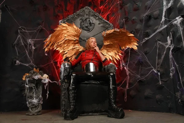 Passionate lazy Cupid blond long hair guy in red leather jacket and vinyl pants with huge Golden wings sits on a huge throne in hell — ストック写真