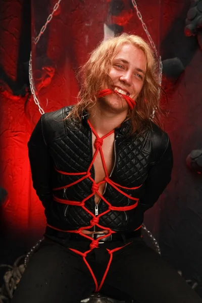 handsome young long-haired guy sits in a leather jacket and is tied with red ropes. sexy bondage slave lobes bdsm and be dominated