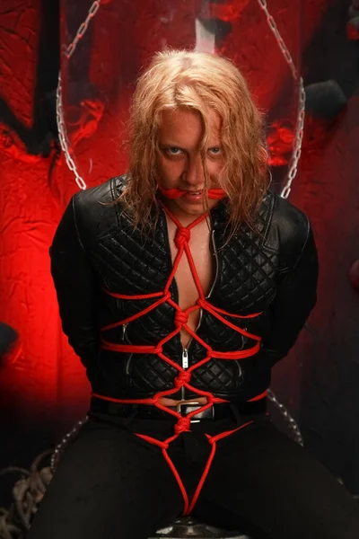 handsome young long-haired guy sits in a leather jacket and is tied with red ropes. sexy bondage slave lobes bdsm and be dominated