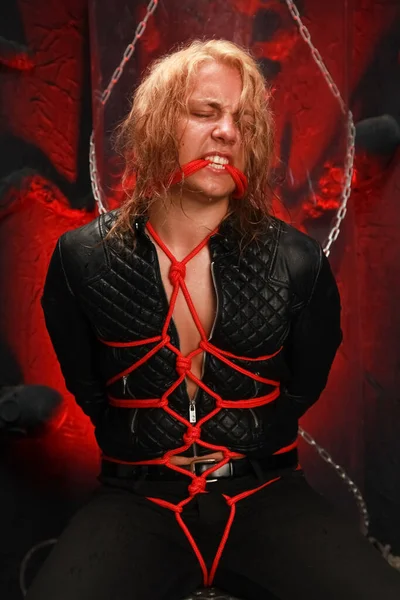 handsome young long-haired guy sits in a leather jacket and is tied with red ropes. sexy bondage slave lobes bdsm and be dominated