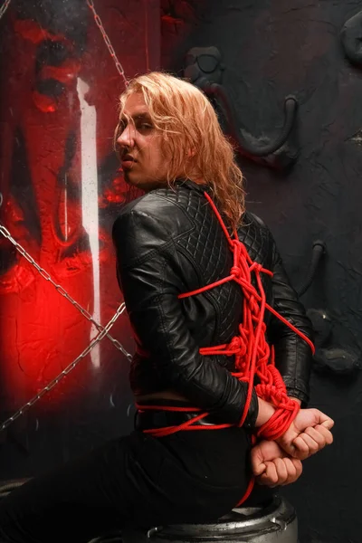 Guapo joven de pelo largo se sienta en una chaqueta de cuero y está atado con cuerdas rojas. sexy esclavitud esclavo lóbulos bdsm y ser dominado — Foto de Stock