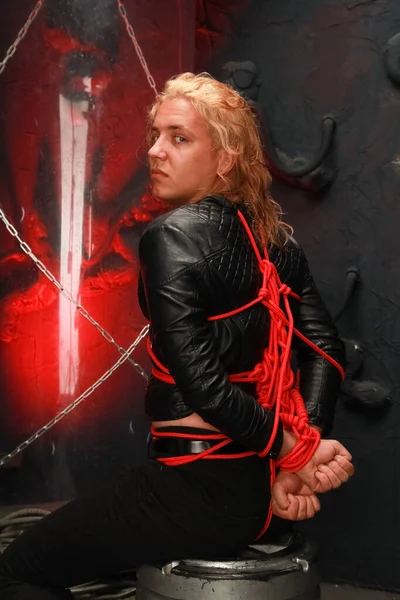 Gutaussehender junger langhaariger Kerl sitzt in einer Lederjacke und ist mit roten Stricken gebunden. sexy Bondage Sklavenlappen bdsm und dominiert werden — Stockfoto