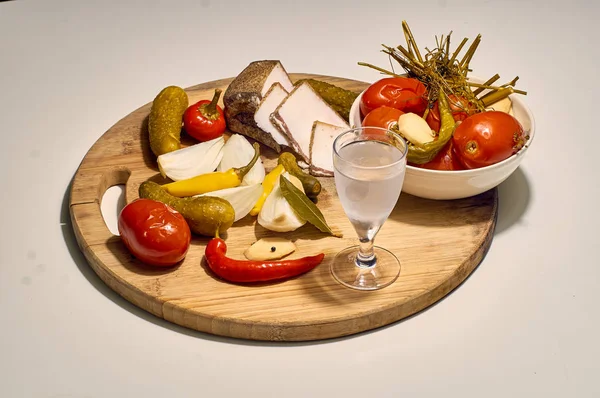 Russian still life: a sweaty glass of icy Russian vodka with a traditional folk snack: lard, pickled cucumbers, hot red pepper, onions and garlic - on a wooden board. Holiday mysterious Russian soul