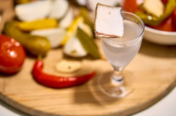 Russian still life: a sweaty glass of icy Russian vodka with a traditional folk snack: lard, pickled cucumbers, hot red pepper, onions and garlic - on a wooden board. Holiday mysterious Russian soul