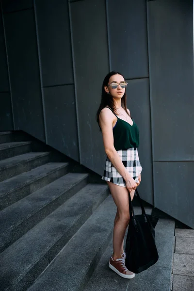 Menina sexy perto de uma parede escura em uma camiseta, óculos de sol e shorts curtos — Fotografia de Stock