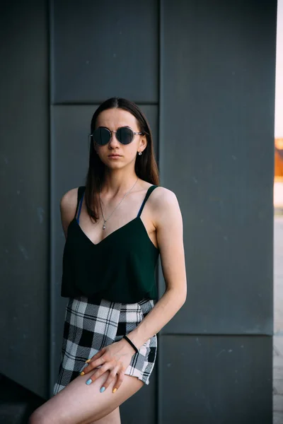 Sexig tjej nära en mörk vägg i en T-shirt, sol glasögon och korta shorts — Stockfoto